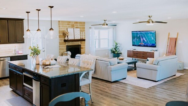 Eine moderne Küche mit Kochinsel, ein Essbereich mit Tisch und Stühlen, ein Wohnzimmer mit Sofa und Fernseher sowie ein Deckenventilator.
