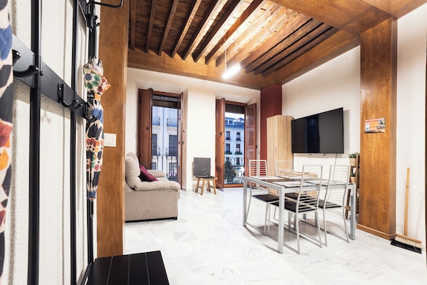 A modern living room with a sofa, dining table, and chairs. There is a TV mounted on the wall and a window with a view of buildings outside.