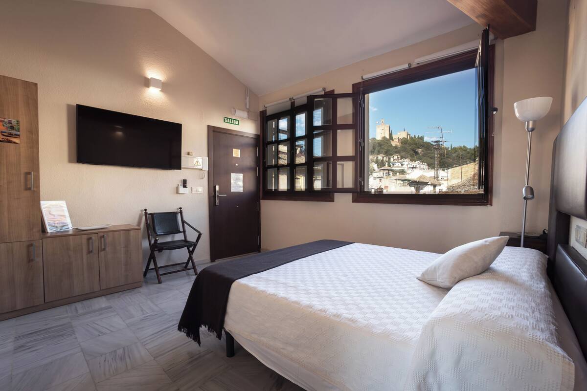 A bedroom with a bed, a desk, a chair, a TV, and a window with a view of a town.