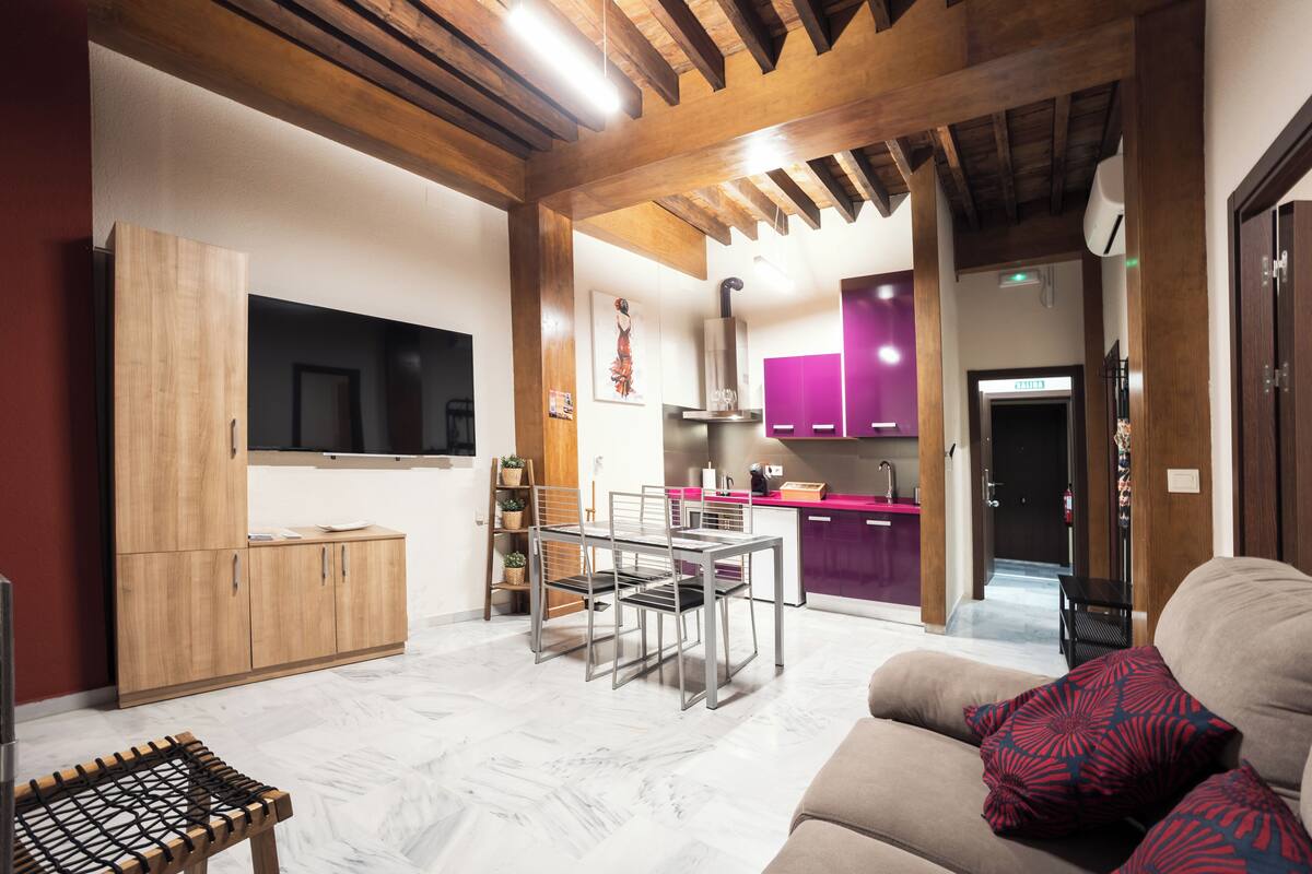 A modern living room with a sofa, a dining table with chairs, a TV mounted on the wall, wooden cabinets, and a kitchen area with purple cabinets and a countertop.