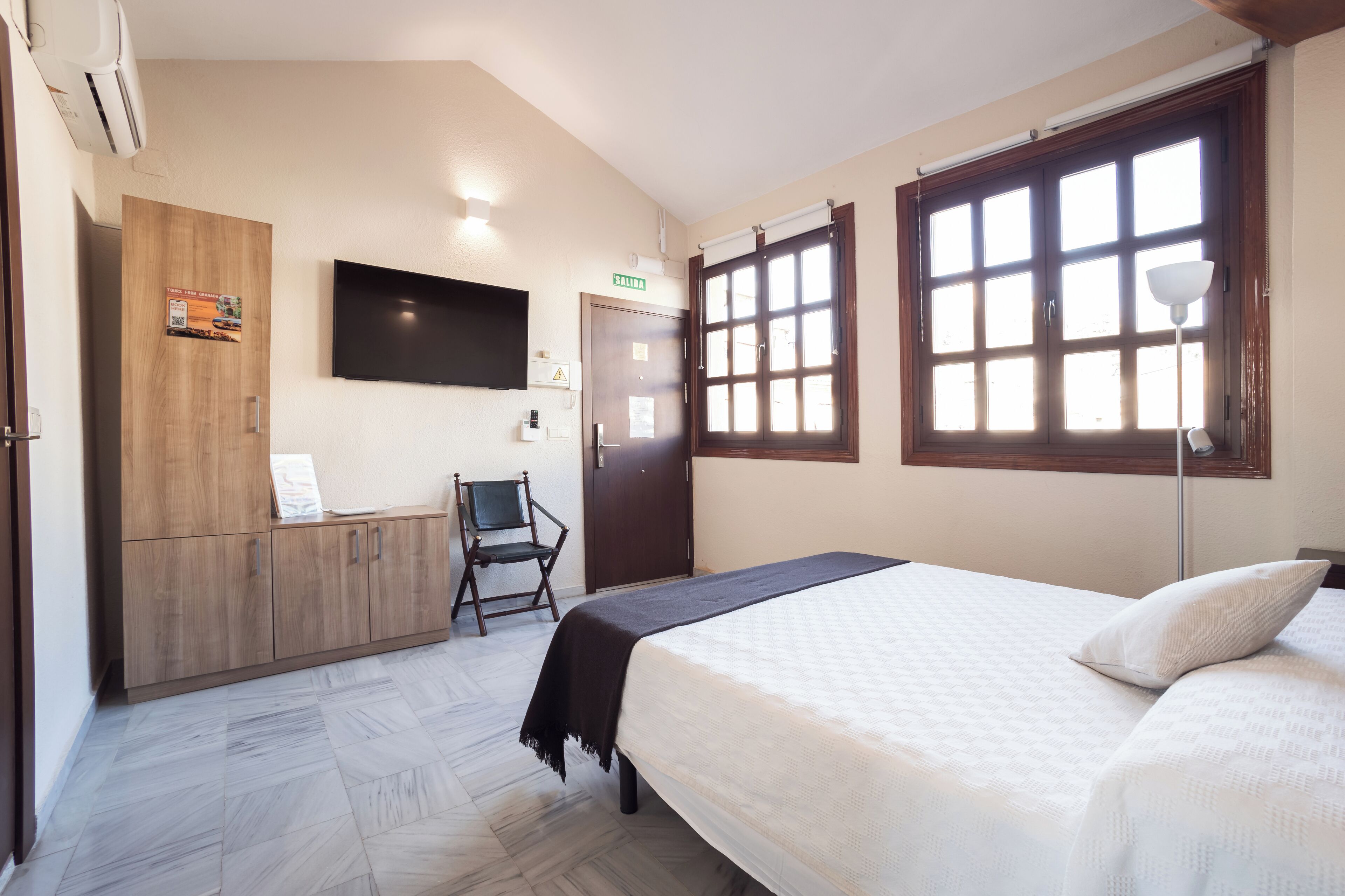A hotel room with a bed, a wooden wardrobe, a chair, a television, and a window with a view.