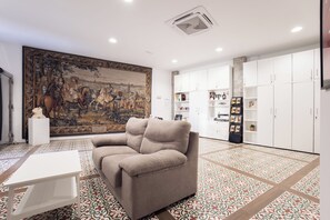 A living room with a beige sofa, a white coffee table, a large tapestry, and white cabinets with shelves. - Apartamentos Reyes Católicos (Granada)