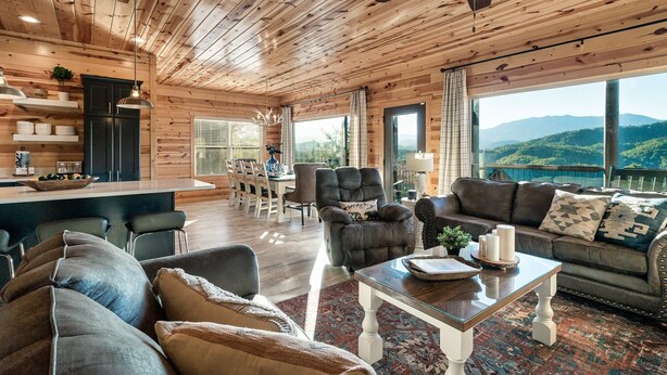 A spacious living room with wooden ceilings, a large sofa, a coffee table, and a view of mountains through large windows.