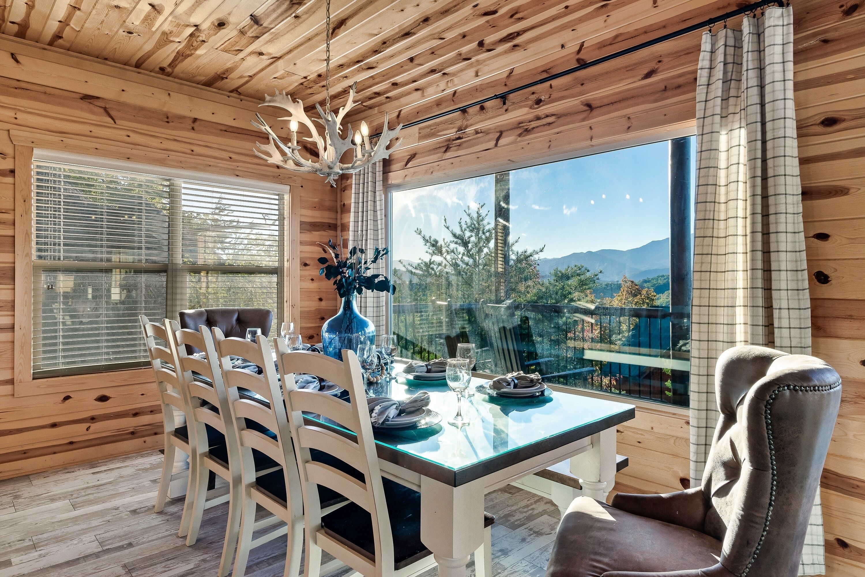 A wooden-paneled room with a dining table set for four, a chandelier, and a large window offering a mountain view.