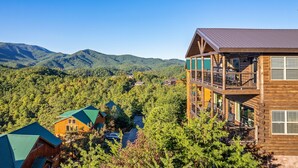 A wooden cabin with a balcony, surrounded by dense forest and mountains in the background.
