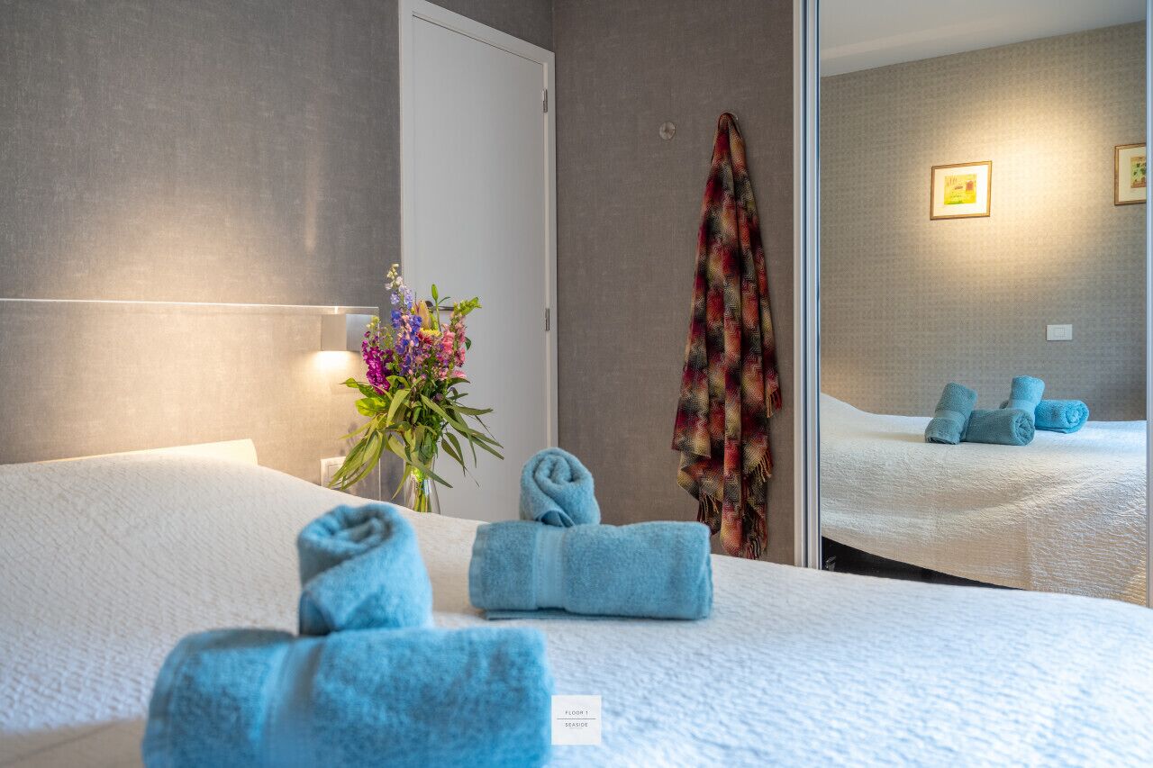 A hotel room with a bed, a mirror, a wall-mounted towel rack, and a flower vase.