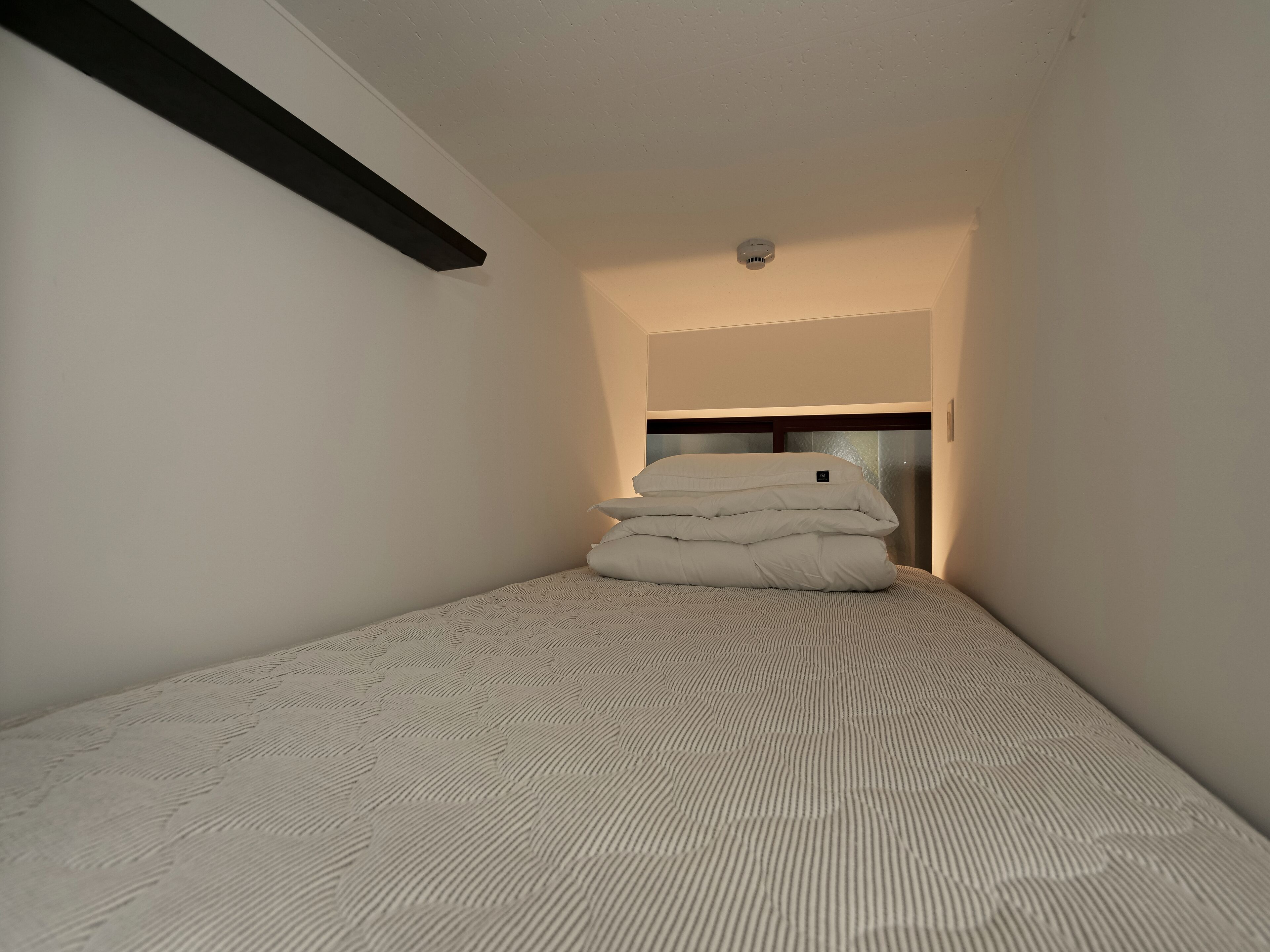 A small, narrow room with a bed at the end, a ceiling light, and a smoke detector.