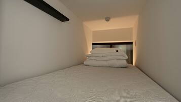 A small, narrow room with a bed at the end, a ceiling light, and a smoke detector.