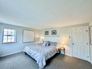 A bedroom with a bed, a nightstand, a lamp, a door, and a window. - Short 1/2 mile walk to the beach! Located in beautiful Harwich. (Harwich)