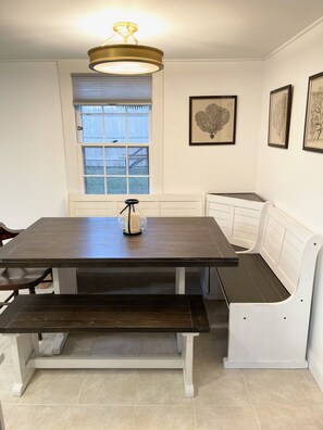 Uma área de refeições com mesa de madeira escura, assentos tipo banco brancos e luminária no teto.