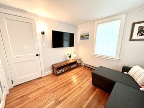 Um quarto com piso de madeira, um sofá cinza, uma TV fixada na parede, uma pequena mesa de madeira com abajur e uma janela com persianas.