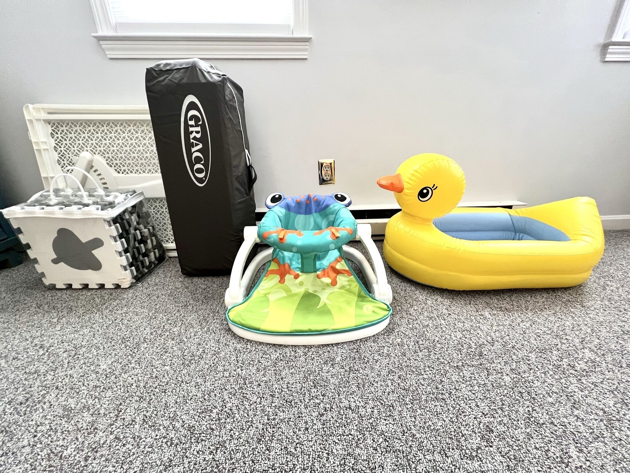 A room with a grey carpet, a white radiator, a black storage bag, a grey storage bin, a blue and green play mat, and a yellow inflatable duck.