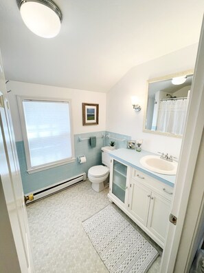 A bathroom with a toilet, sink, and mirror. There is a window with blinds, a picture on the wall, and a towel rack. - Short 1/2 mile walk to the beach! Located in beautiful Harwich. (Harwich)