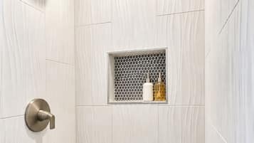 A shower with a rain showerhead, a wall-mounted soap dispenser, and a small shelf with two bottles.