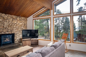 Un salon chaleureux avec une cheminée en pierre, un téléviseur et une grande fenêtre donnant sur la forêt.