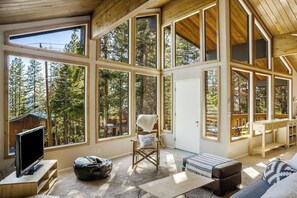 A spacious room with large windows offering a view of a forest. There is a television on a wooden stand, a chair with a towel, and a sofa with a patterned cushion.