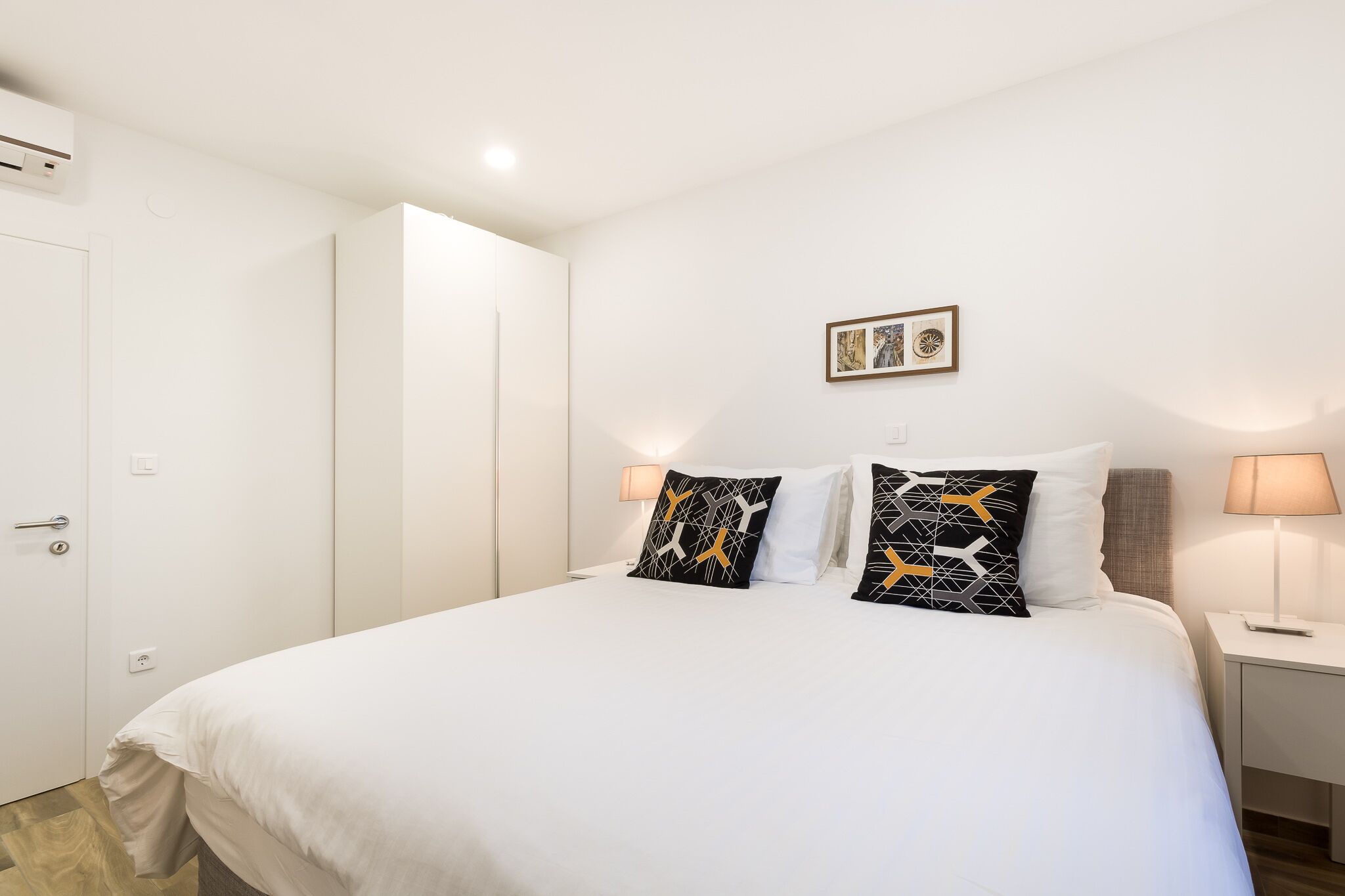 Una habitación de hotel con una cama grande, dos almohadas, una mesita de noche con una lámpara, un cuadro en la pared y una puerta con manija.