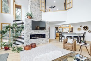 Amplia sala de estar con chimenea de piedra, televisor de pantalla plana y comedor, con una cocina al fondo.