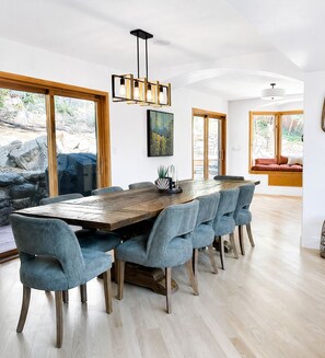 A modern dining area with a large wooden table, blue upholstered chairs, and a hanging light fixture. - 6BR Mountain Retreat • Hot Tub • Walk to Pearl St (Boulder)