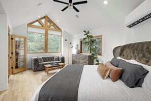 Un dormitorio con una cama grande, un sofá, piso de madera y vista a árboles por las ventanas.