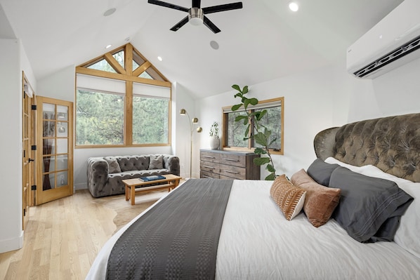 A bedroom with a large bed, a sofa, a wooden floor, and a view of trees through the windows.