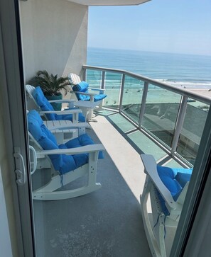 A balcony with white rocking chairs and blue cushions, overlooking a beach with clear blue water and a distant shoreline.