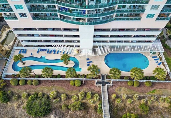 A multi-story hotel with a pool area, including a large pool and a smaller pool, surrounded by palm trees and lounge chairs.