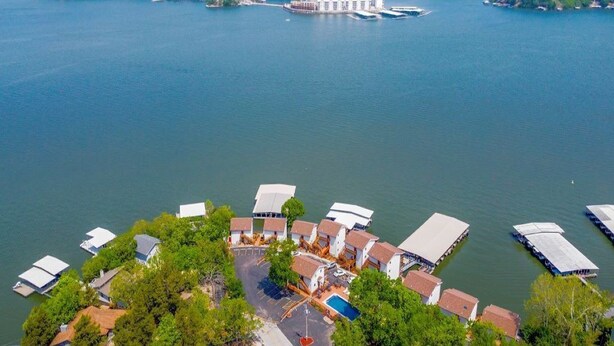 Une vue aérienne d’un hôtel situé au bord d’un lac, avec plusieurs bateaux amarrés, un bâtiment principal doté d’une haute cheminée et un parking entouré d’arbres.