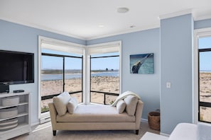 A room with a large window, a beige sofa, a flat-screen TV, and a beach view.