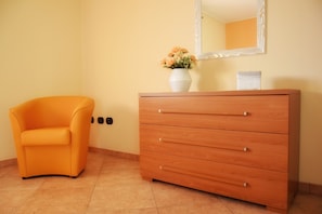 Una habitación con un aparador de madera, un jarrón blanco con flores y una silla naranja.