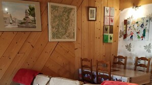 A cozy room with a wooden floor, a sofa with patterned cushions, a dining table with chairs, and various framed artworks on the walls. - ALL WOODEN APARTMENT WITH GARDEN 100 meters from town center in quiet location. (Gérardmer)