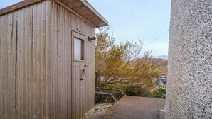 Un capanno di legno con una porta e una finestra, adiacente a un muro in pietra e un vialetto sterrato.