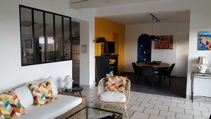Ein Wohnzimmer mit einer Couch, einem Sessel, einem Esstisch und einer Küchenzeile mit Fernseher und Schränken.