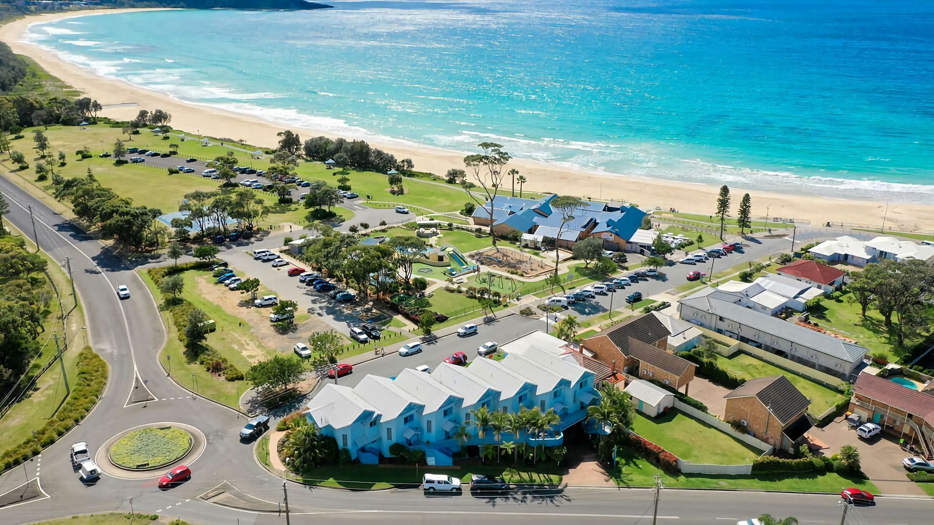 An aerial view of a coastal area with a beach, a parking lot, and buildings with blue roofs.