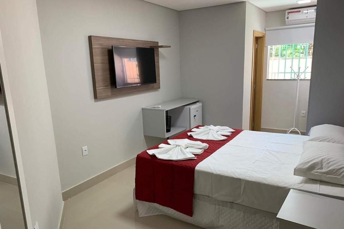 A hotel room with a bed, a TV mounted on the wall, a small desk with a chair, and a window with a view of greenery.