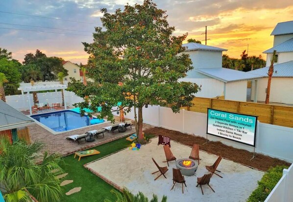 A backyard with a swimming pool, a fire pit, and a sign advertising Coral Sands with amenities like a gas-heated pool, separate kitchen, tiki bar, and beach access.