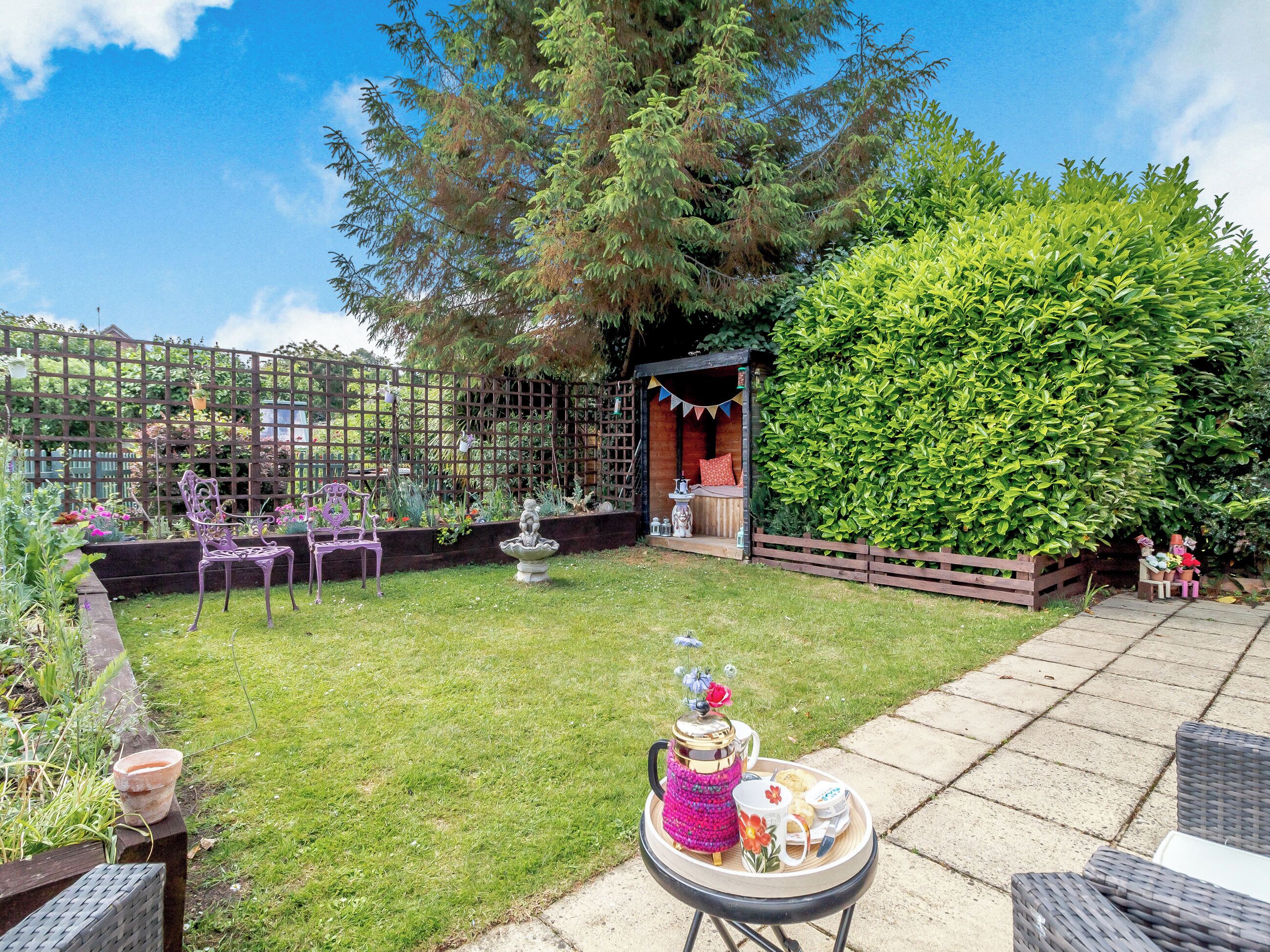 A backyard with a lawn, a wooden deck, a seating area with chairs, and a small structure with a roof.