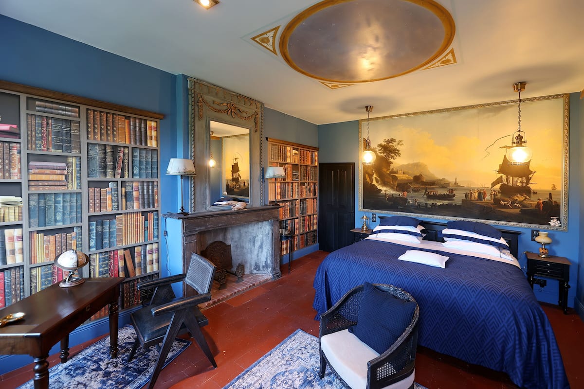 A bedroom with a large bed, a fireplace, a bookshelf, and a large painting on the wall.