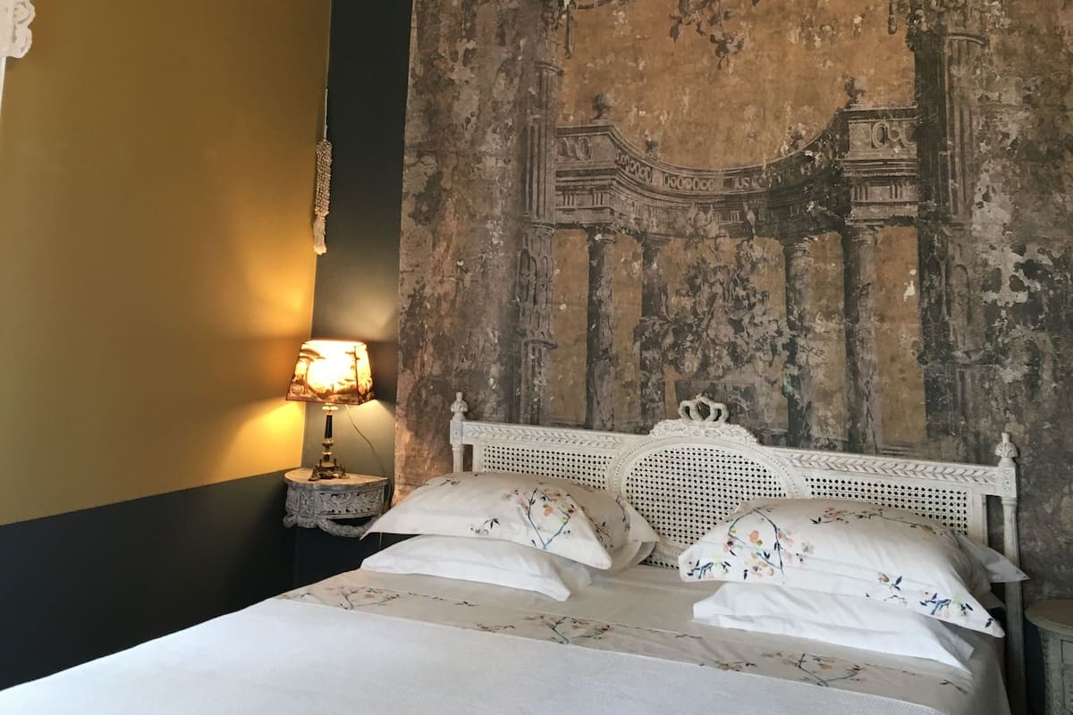 A bed with a white headboard, a wall with a mural, a hanging light fixture, and a nightstand with a lamp.