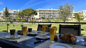 Mesa de café da manhã com pratos, xícaras e croissants numa varanda com vista para uma piscina e edifícios de hotel.