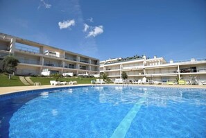 Piscina com espreguiçadeiras, um edifício com varandas e céu azul.