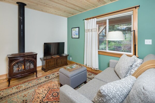 A cozy living room with a wooden floor, a fireplace, a television, and a large window with curtains.