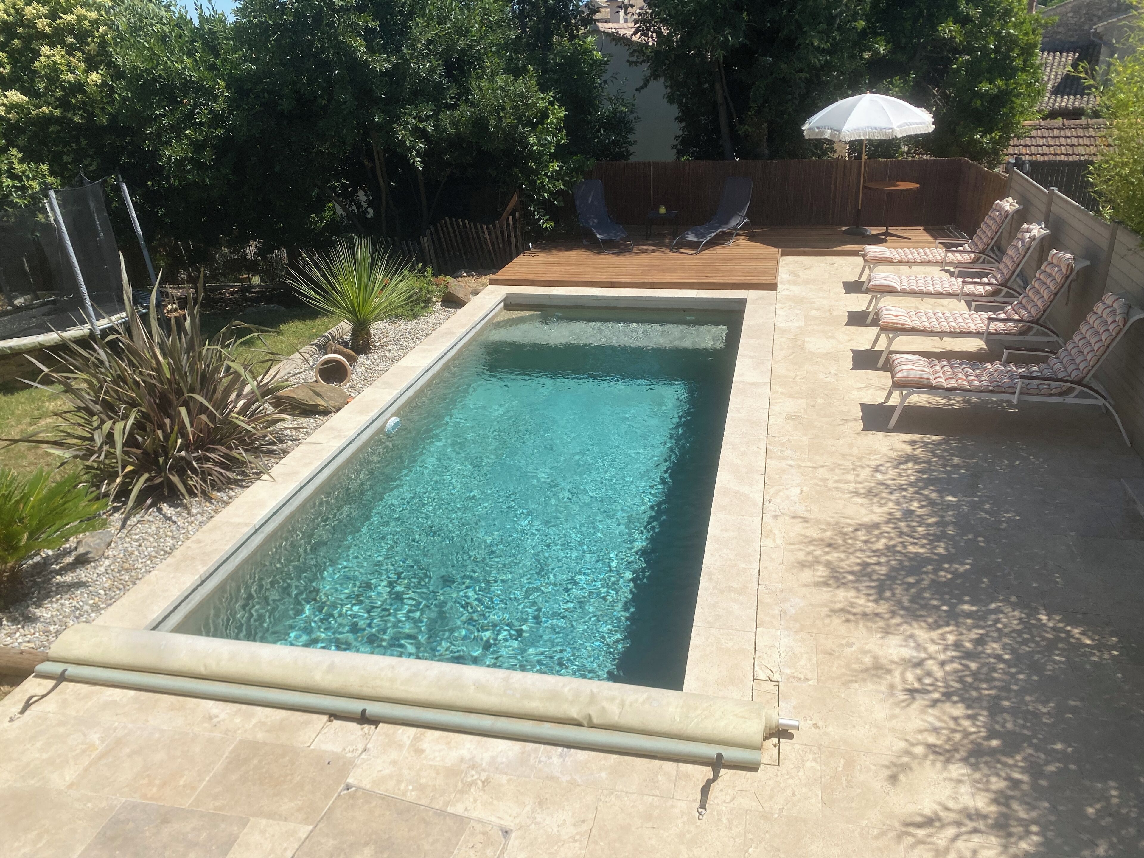 Ein rechteckiger Pool mit klarem Wasser, umgeben von einer gefliesten Liegefläche. Liegestühle und ein kleiner Tisch mit Sonnenschirm sind vorhanden.