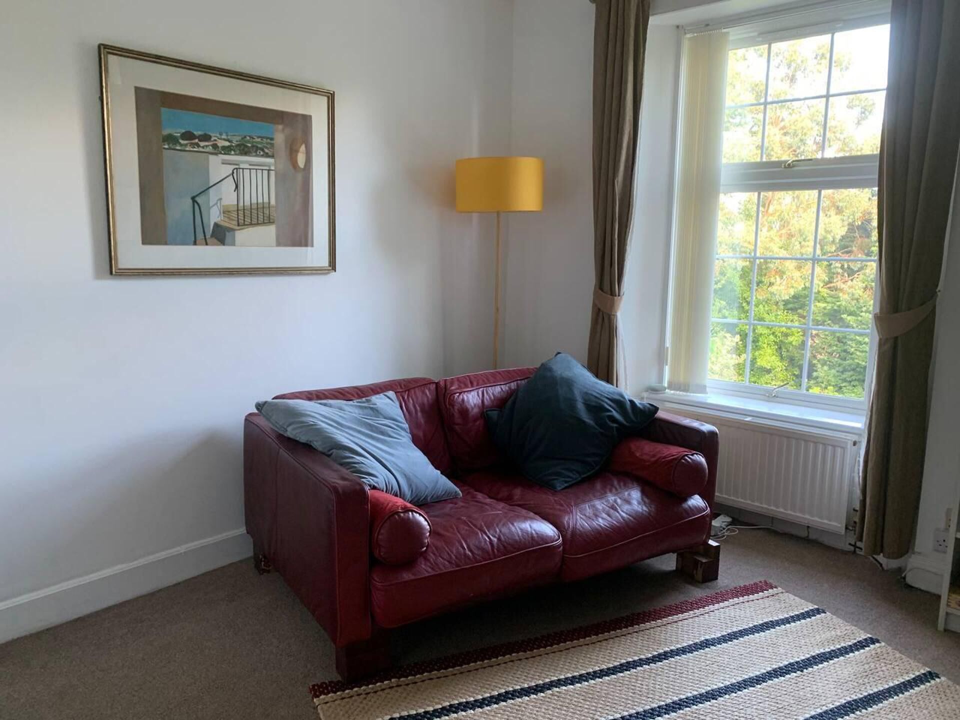 A red leather sofa with two pillows, a framed picture on the wall, a floor lamp, and a window with curtains.