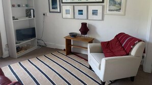 Uma sala de estar com uma poltrona branca, um tapete listrado, uma estante de livros e obras de arte emolduradas na parede.