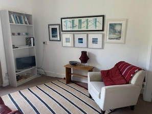 A living room with a white armchair, a striped rug, a bookshelf, and framed artwork on the wall. - Cedarbank's - The Little Art House - dog Friendly (Dunoon)