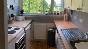 A compact kitchen with a window, a stove, a sink, and a trash bin. - Cedarbank's - The Little Art House - dog Friendly (Dunoon)