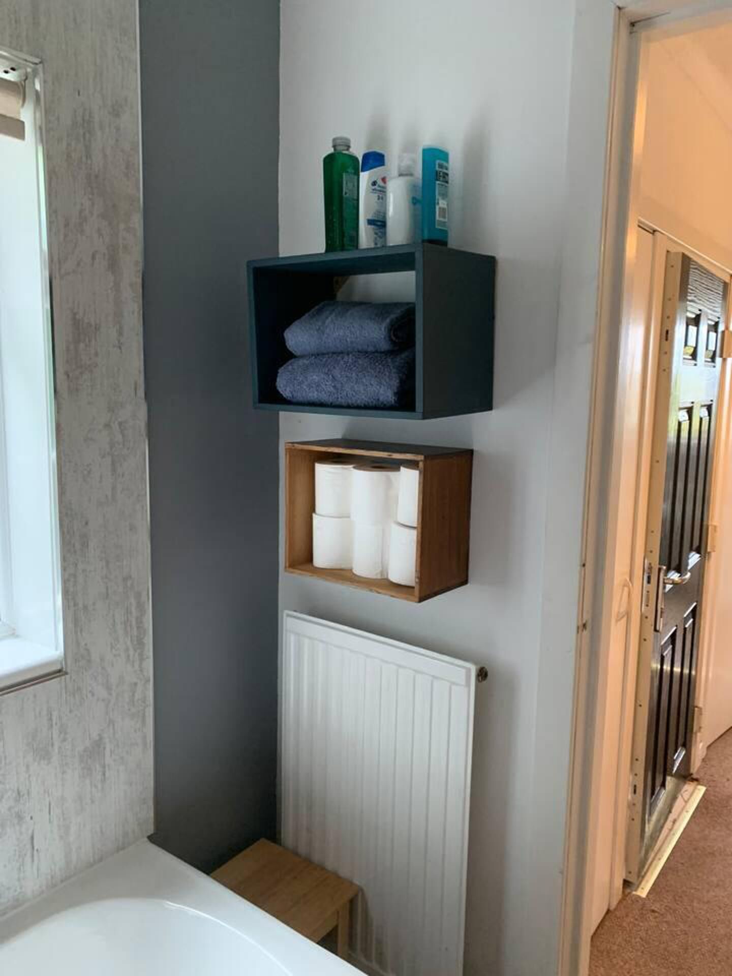A bathroom with a white bathtub, a wooden stool, a radiator, and shelves with toiletries.