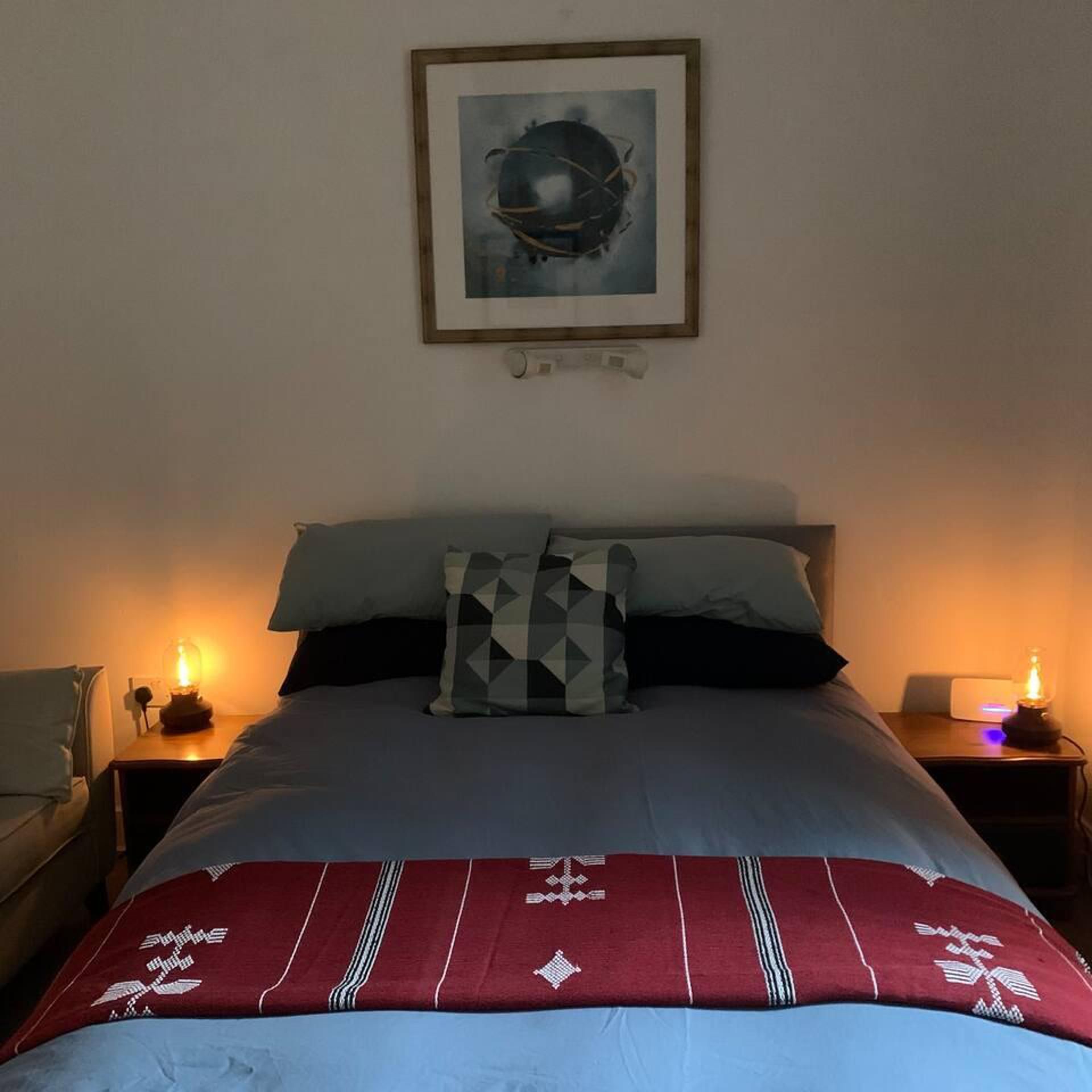 Quarto com uma cama, duas luminárias de apoio, um quadro na parede e um cobertor com padrão vermelho e branco.