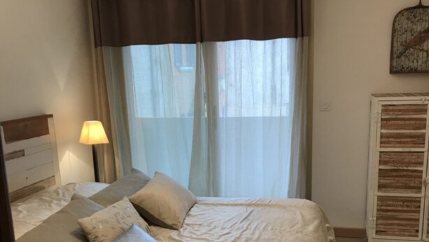 A neatly made bed with a headboard, a bedside lamp, and a window with sheer curtains.